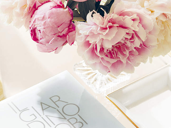 Auf einem Tisch steht eine Kristallvase mit rosa und weißen Pfingstrosen. Daneben steht ein Buch mit dem Titel „IL GIARDINO“ und eine weiße Schale mit Goldrand. Die Szene ist hell und elegant.
