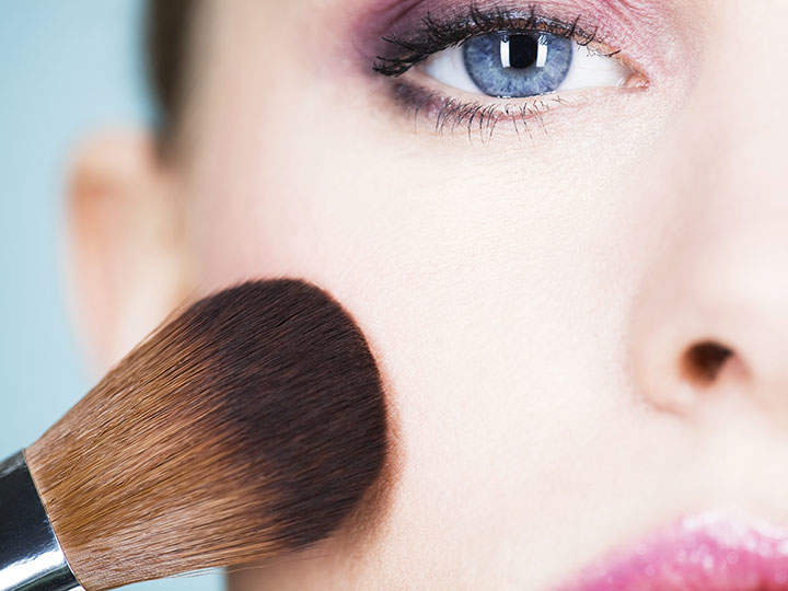 Nahaufnahme einer Person, die mit einem Make-up-Pinsel Rouge auf ihre linke Wange aufträgt. Das mit violettem Lidschatten hervorgehobene Auge ist sichtbar und die Haut erscheint glatt. Das Bild konzentriert sich auf die Technik des Schminkens.
