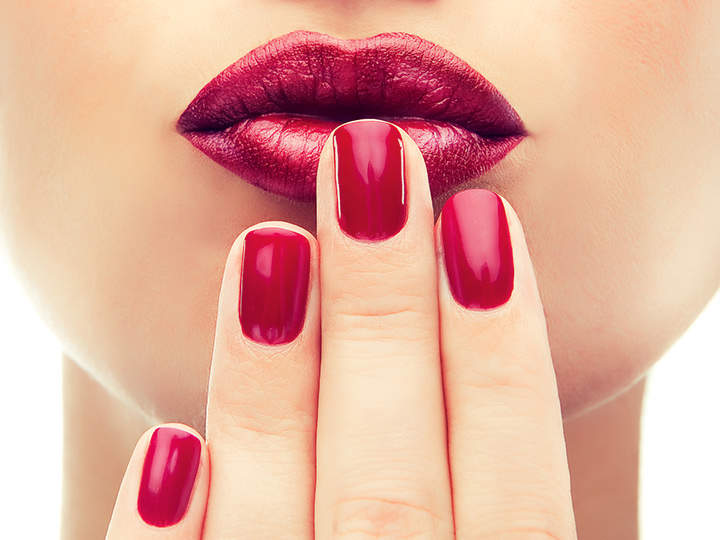 Nahaufnahme einer Person mit leuchtend rotem Lippenstift und passendem rotem Nagellack, die ihre Lippen sanft mit manikürten Fingern berührt. Das Bild betont die kräftigen, glänzenden Farben und die glatte Textur auf Lippen und Nägeln.