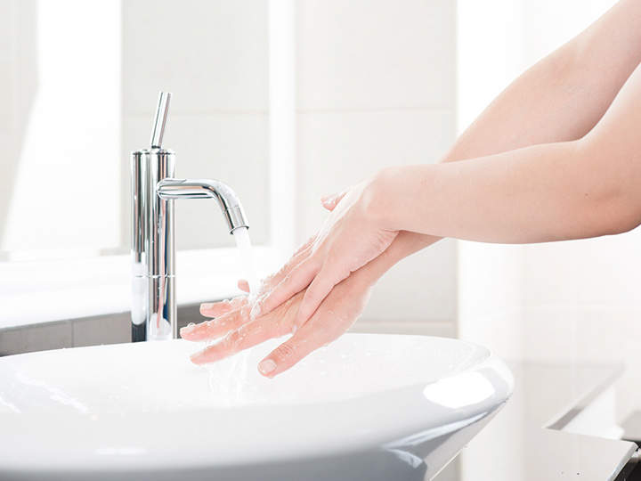 Eine Person wäscht sich die Hände in einem weißen Waschbecken mit einem modernen Chromhahn. Das Wasser läuft und Seifenschaum bedeckt die Hände. Der Hintergrund ist sanft beleuchtet und schafft eine saubere und helle Atmosphäre.