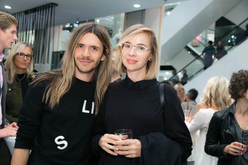 Ein Mann mit langen Haaren und eine Frau mit kurzen blonden Haaren und Brille stehen zusammen, lächeln und halten ein Getränk in der Hand. Sie sind bei einer gesellschaftlichen Veranstaltung in einem Gebäude, umgeben von anderen Menschen, mit Treppen und modernem Dekor im Hintergrund.