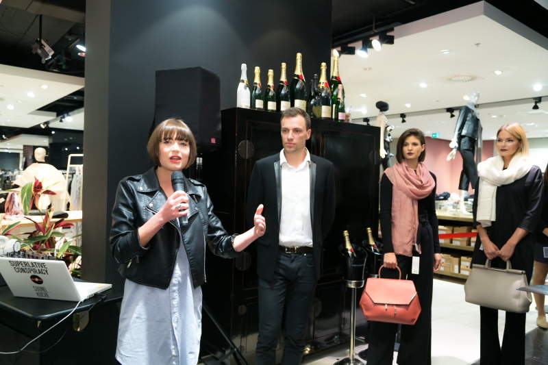 Eine Person mit einem Mikrofon spricht zu einem Publikum in einem schicken Geschäft. Drei weitere Personen stehen in der Nähe, im Hintergrund sind auf einem Schrank Champagnerflaschen ausgestellt. Sie scheinen an einer Mode- oder Einzelhandelsveranstaltung teilzunehmen.