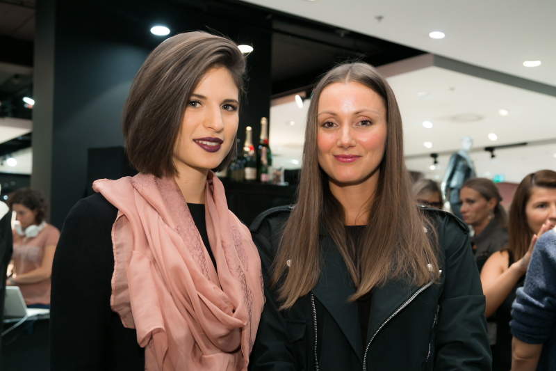 Zwei Frauen stehen zusammen in einem Innenraum. Die Frau links hat kurzes braunes Haar und trägt einen rosa Schal. Die Frau rechts hat langes braunes Haar und trägt eine schwarze Jacke. Im Hintergrund sind weitere Personen und Flaschen zu sehen.