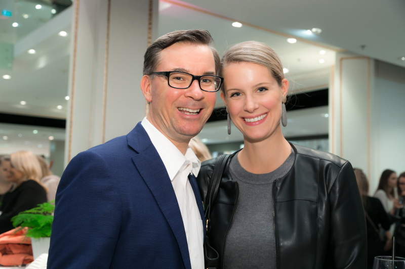 Ein Mann in blauem Anzug und Brille steht neben einer Frau mit Pferdeschwanz, die eine schwarze Jacke über einem grauen Oberteil trägt. Sie lächeln in die Kamera in einem hell erleuchteten Innenraum.