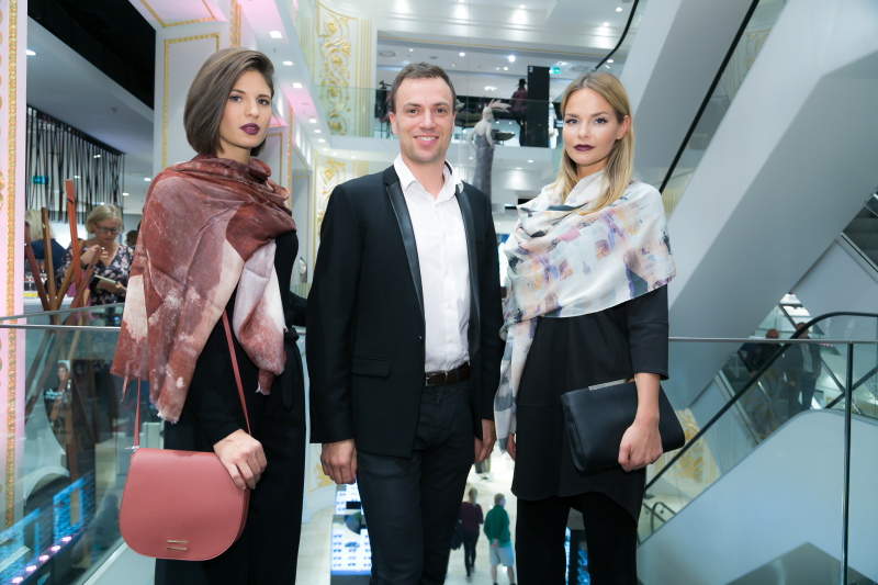Ein Mann im Anzug steht zwischen zwei Frauen, die schicke Schals und schwarze Outfits tragen. Sie posieren in einem modernen mehrstöckigen Einzelhandelsgeschäft mit Rolltreppen und heller Beleuchtung im Hintergrund.