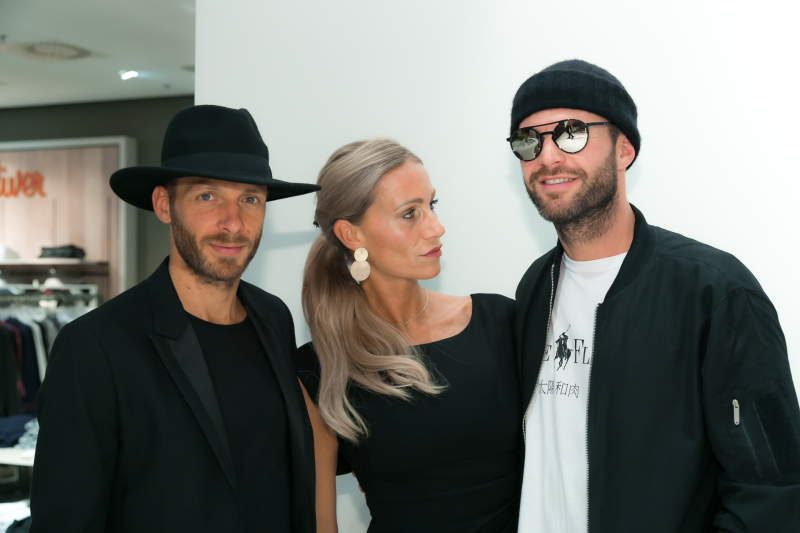 Drei Personen in schicken schwarzen Outfits stehen drinnen zusammen. Der Mann links trägt einen Fedora, die Frau in der Mitte hat lange blonde Haare und große Ohrringe und der Mann rechts trägt eine Sonnenbrille und eine Beanie.