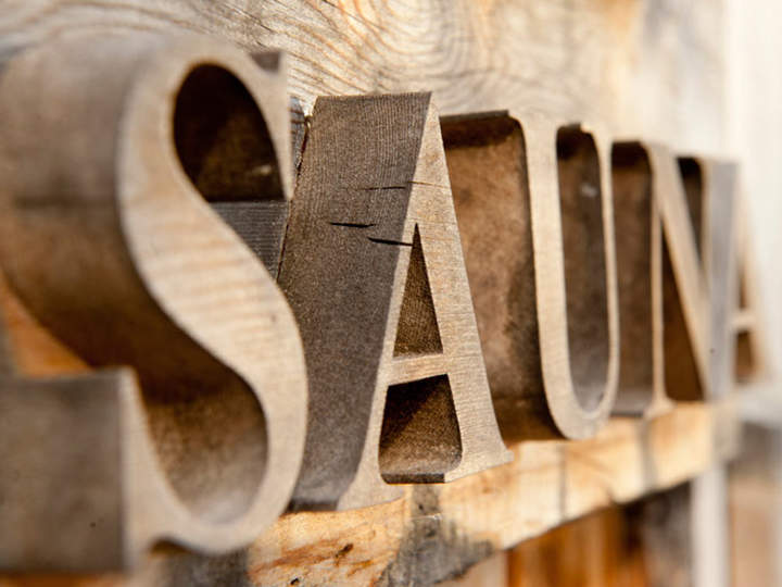 Das Bild zeigt große Holzbuchstaben mit der Aufschrift „SAUNA“, die auf einer rustikalen Holzoberfläche angebracht sind. Die Nahaufnahme hebt die Textur des Holzes und die Maserung der Buchstaben und des Hintergrunds hervor.
