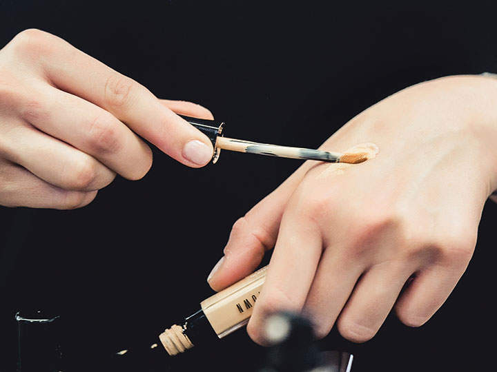 Eine Person trägt Concealer mit einem Applikatorstab auf den Handrücken auf. Der Concealer befindet sich in einer kleinen Tube, wobei das Etikett teilweise sichtbar ist. Der Hintergrund ist dunkel und hebt die Hand und das Make-up-Produkt hervor.