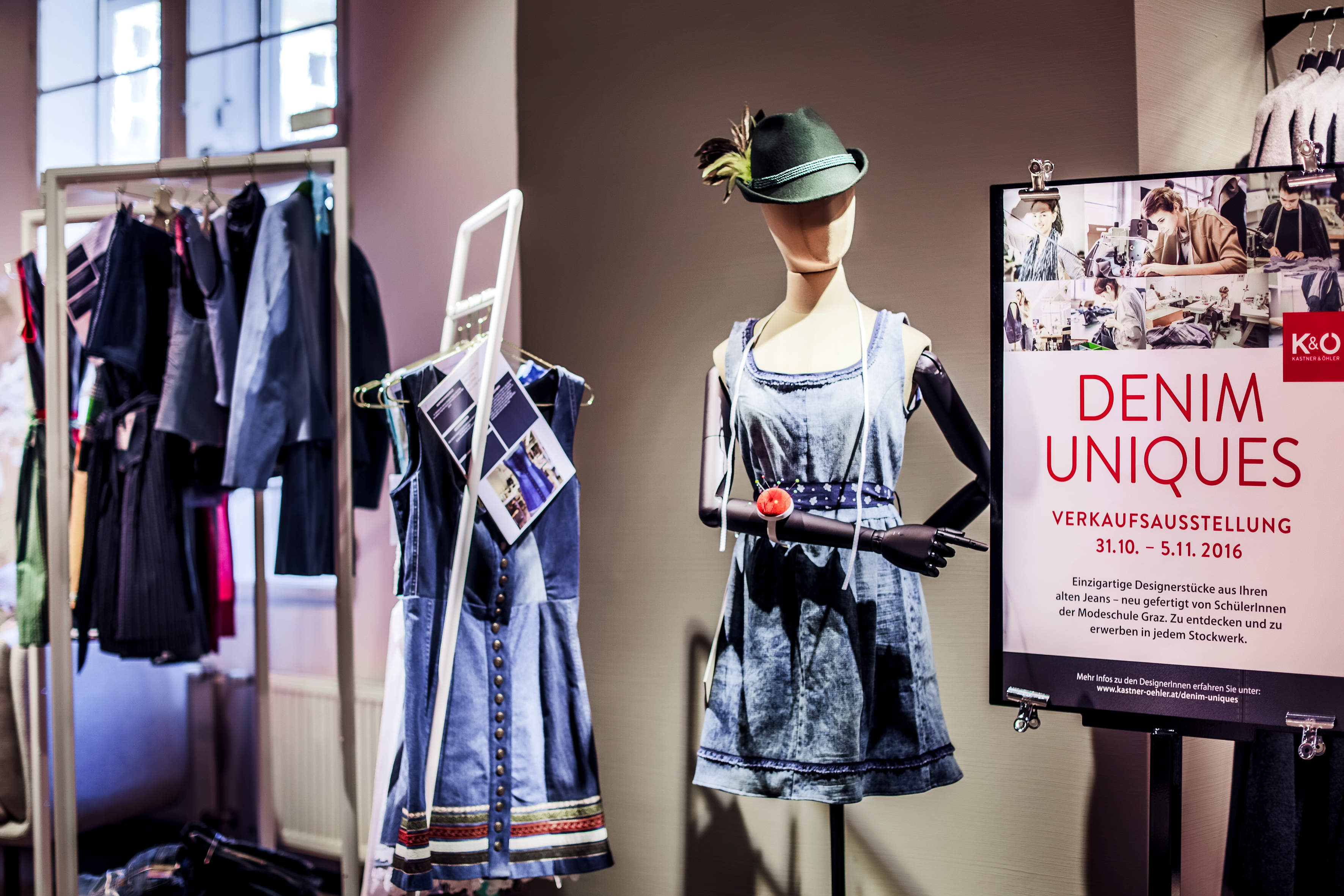 Eine stilvolle Präsentation von Outfits mit Jeansmotiven in einer Modeausstellung. Eine Schaufensterpuppe trägt ein Jeanskleid und einen Hut neben einem Plakat, das die Veranstaltung „Denim Uniques“ vom 31. Oktober bis 5. November 2016 ankündigt. Kleiderständer mit weiteren Kleidungsstücken im Hintergrund.