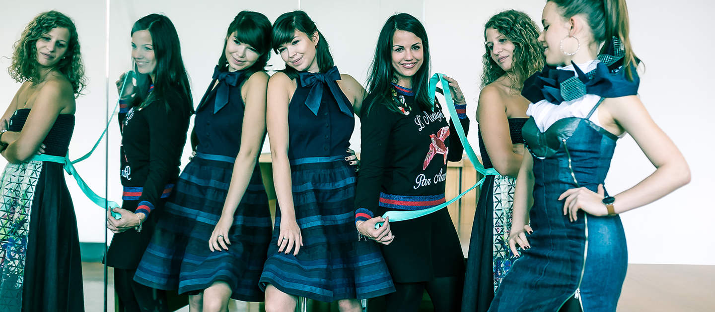 Eine Gruppe von Frauen in aufeinander abgestimmten blauen Outfits steht zusammen, einige von ihnen blicken vor einen Spiegel, sodass sich Reflexionen bilden. Sie posieren in einem hellen Raum und präsentieren ihre stilvolle Kleidung.