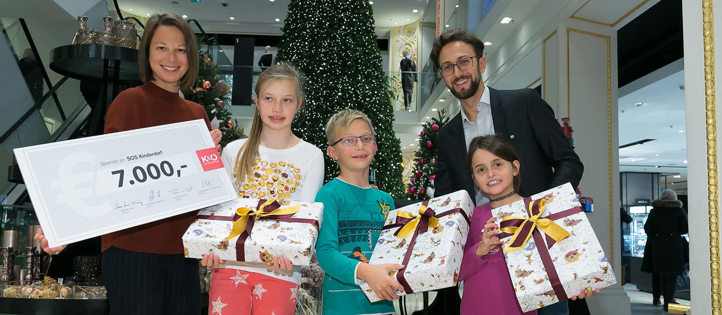 Eine Gruppe aus zwei Erwachsenen und drei Kindern steht in einem geschmückten Raum mit einem Weihnachtsbaum. Die Erwachsenen halten einen großen Scheck über 7.000 Euro und die Kinder verpackte Geschenke mit Schleifen. Sie lächeln alle.