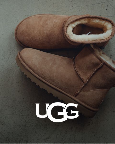 Ein Paar brauner UGG-Wildlederstiefel mit Plüsch-Innenfutter, das auf einer grauen Oberfläche ruht. Das UGG-Logo ist am unteren Rand des Bildes gut sichtbar angebracht.