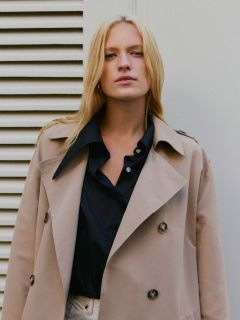 Eine Person mit langen blonden Haaren trägt einen beigen Trenchcoat über einem schwarzen Hemd und steht vor einer weißen Lamellenwand. Sie hat einen neutralen Gesichtsausdruck.