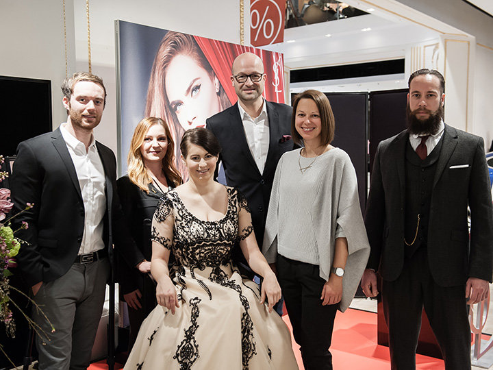 Eine Gruppe von sechs Personen, drei Männer und drei Frauen, steht lächelnd im Innenbereich. In der Mitte der Gruppe sitzt eine Frau in Abendkleid. Sie sind elegant gekleidet und scheinen sich auf einer Veranstaltung oder Versammlung zu befinden.