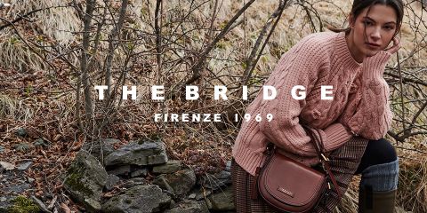 Eine Frau sitzt draußen in einer natürlichen Umgebung. Sie trägt einen grob gestrickten rosa Pullover und hält eine braune Lederhandtasche in der Hand. Der Text „The Bridge Firenze 1969“ ist über das Bild gelegt. Im Hintergrund sind trockenes Gras und Felsen zu sehen.