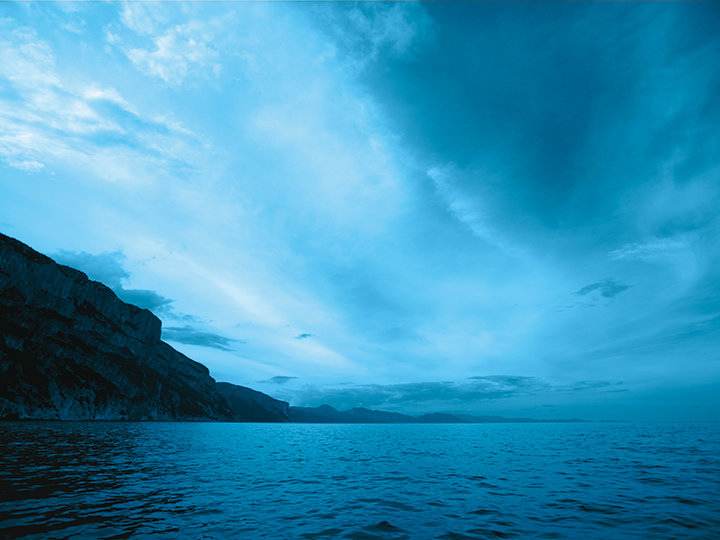 Eine heitere Meereslandschaft mit ruhigem, blauem Wasser, das sich bis zu den entfernten Berglandschaften erstreckt, unter einem weiten Himmel voller weicher, dünner Wolken. Die Szene ist in Blautöne getaucht, was eine ruhige und friedliche Atmosphäre schafft.