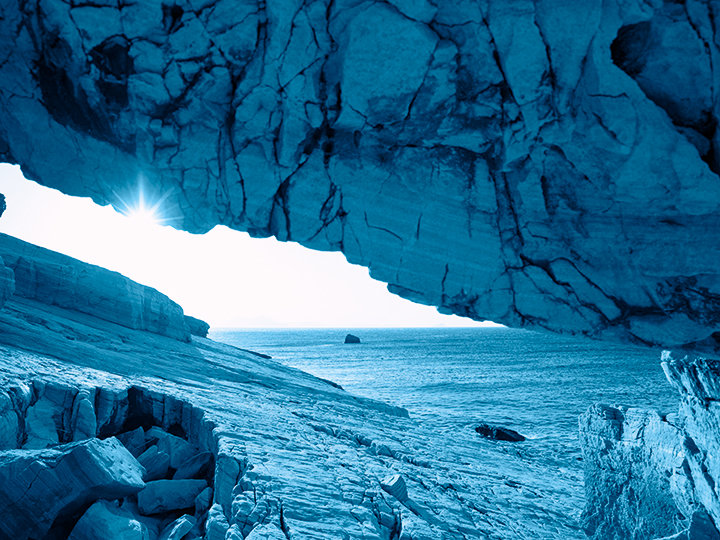 Eine Küstenhöhle mit schroffen Felsformationen überblickt das Meer. Sonnenlicht fällt durch die Öffnung, beleuchtet den felsigen Vordergrund und schafft eine ruhige, azurblaue Landschaft aus Meer und Himmel.