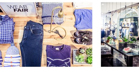 Auf einem Holztisch liegen gefaltete blaue und gestreifte Kleidungsstücke, Jeans und Schuhe. Auf einem Schild steht „WEAR FAIR“. Daneben steht ein schlichter schwarzer Tisch mit grünen Pflanzen und einem dekorativen Buch, über dem Glühbirnen hängen.