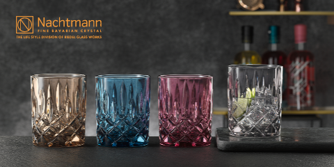 Auf einer dunklen Oberfläche sind vier Kristallgläser in Braun, Blau, Lila und Klar zu sehen. Das Klarglas enthält Eis. In der oberen linken Ecke ist ein Logo für Nachtmann Fine Bavarian Crystal zu sehen. Im Hintergrund sind Flaschen verschwommen.