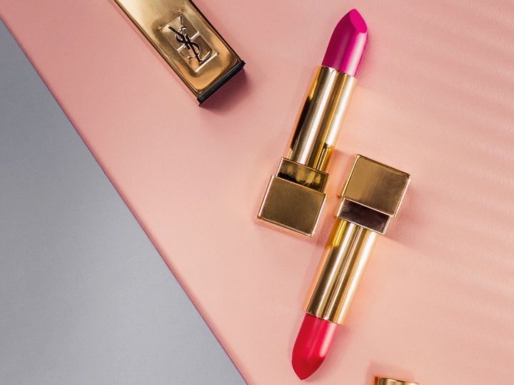Zwei Lippenstifte mit goldenem Gehäuse auf einem diagonal geteilten Hintergrund in zartem Rosa und Grau. Ein Lippenstift ist leuchtend rosa, der andere rot, mit einer geöffneten Kappe daneben. Luxuriöse und elegante Präsentation.