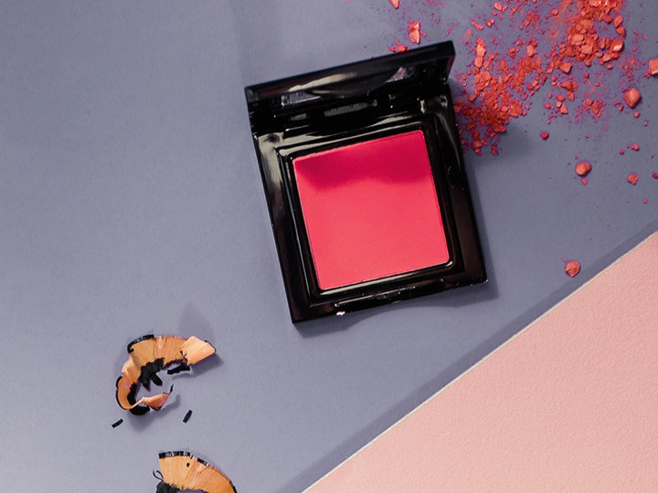 Auf einem pastellfarbenen Hintergrund ist eine rosa Rouge-Dose geöffnet, umgeben von Bleistiftspänen und verstreutem Rougepuder.