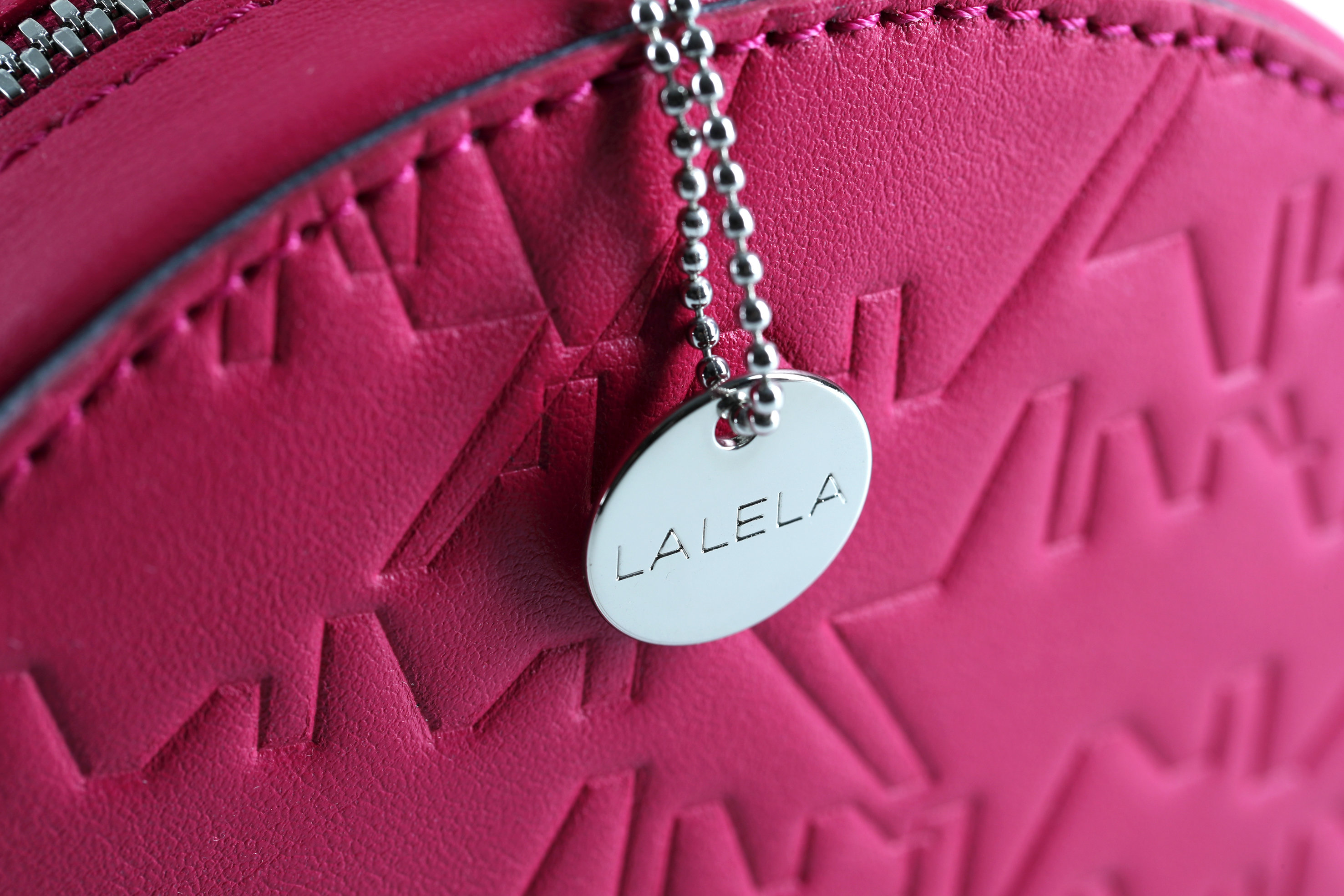 Nahaufnahme einer strukturierten rosa Tasche mit einem runden Metallanhänger mit der Gravur „LALELA“, der an einer Kette befestigt ist. Die Tasche weist ein Zickzack-Prägemuster und einen sichtbaren Reißverschluss an der Oberseite auf.