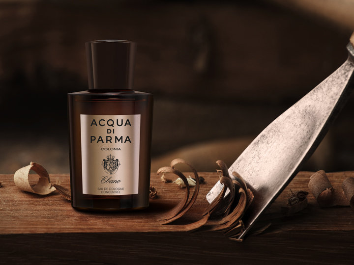 Eine Flasche Acqua di Parma Colonia Cologne steht auf einer Holzoberfläche, umgeben von Holzspänen. In der Nähe steht ein Schnitzwerkzeug, was eine rustikale und handwerkliche Atmosphäre schafft.
