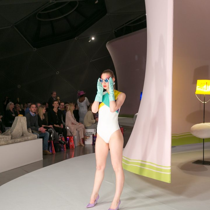 Ein Model in einem weißen Badeanzug und blauen Handschuhen posiert auf einem futuristischen Laufsteg. Im Hintergrund sieht das Publikum zu. Die Kulisse besteht aus großen geometrischen Strukturen und einer modernen Lampe.