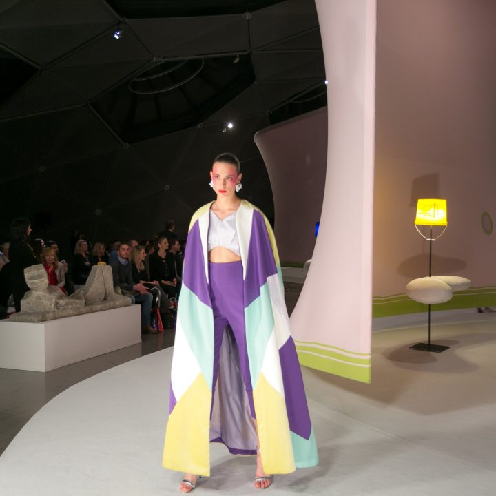 Ein Model läuft in einem farbenfrohen, fließenden Outfit mit einem geometrischen Muster in Lila, Gelb und Blau über den Laufsteg. Im Hintergrund ist eine moderne, künstlerische Installation mit geschwungenen Formen zu sehen. Das Publikum schaut in dem schwach beleuchteten Raum zu.