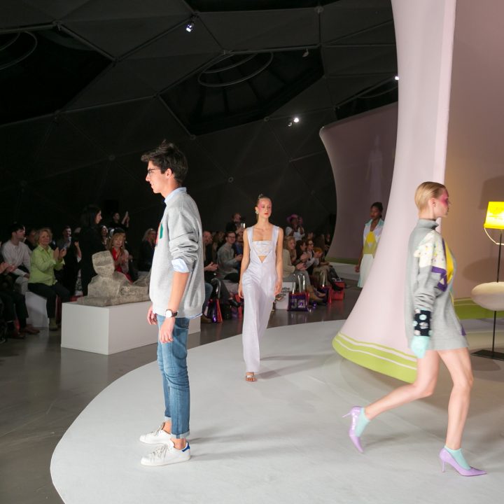 Models laufen in stylischen Outfits über den Laufsteg, darunter ein Mann in lässigem Pullover und Jeans und Frauen in eleganten Kleidern. Das Publikum schaut aufmerksam von der Seite in einem modernen Ambiente mit sanfter Beleuchtung zu.