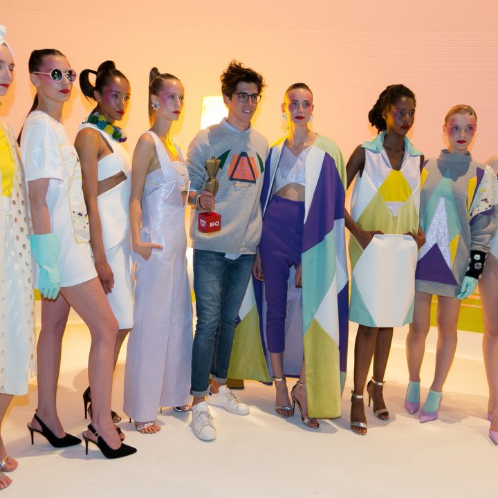 Eine Gruppe von Models präsentiert neben einer leger gekleideten Person farbenfrohe, geometrische Modedesigns. Die Outfits zeichnen sich durch kräftige Formen und leuchtende Farben aus. Die Models und die Person stehen vor einem rosa Hintergrund.