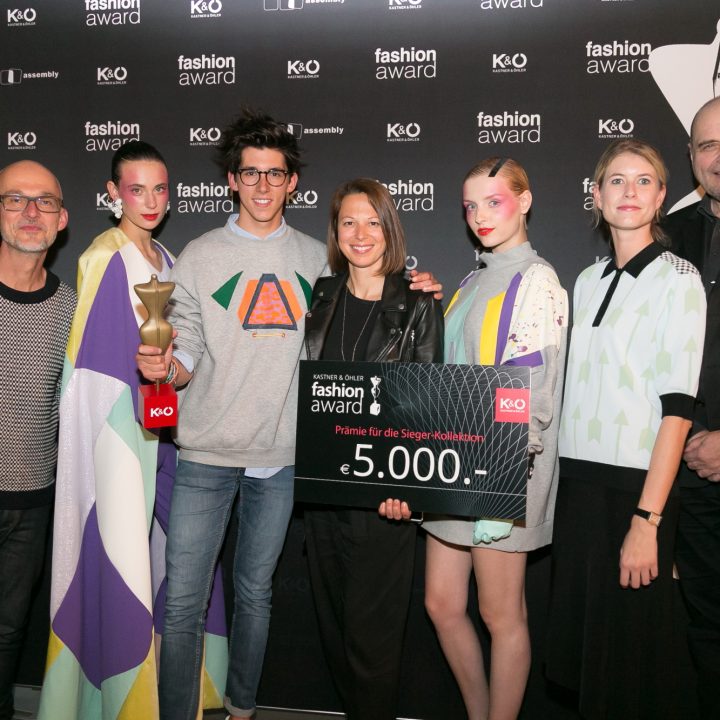 Eine Gruppe von sieben Personen lächelt vor einem schwarzen Hintergrund mit „Fashion Award“-Logos. Eine Person hält eine Trophäe in der Hand, eine andere einen großen Scheck über 5.000 Euro. Sie sind alle in stilvolle, farbenfrohe Kleidung gekleidet.