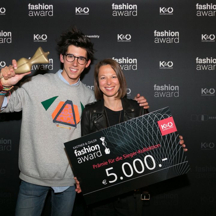 Zwei lächelnde Personen posieren bei einer Fashion Award-Veranstaltung. Einer hält eine Trophäe, der andere einen großen Scheck über 5.000 Euro. Beide stehen vor einem Hintergrund mit den Logos „Fashion Award“ und „K&O“.