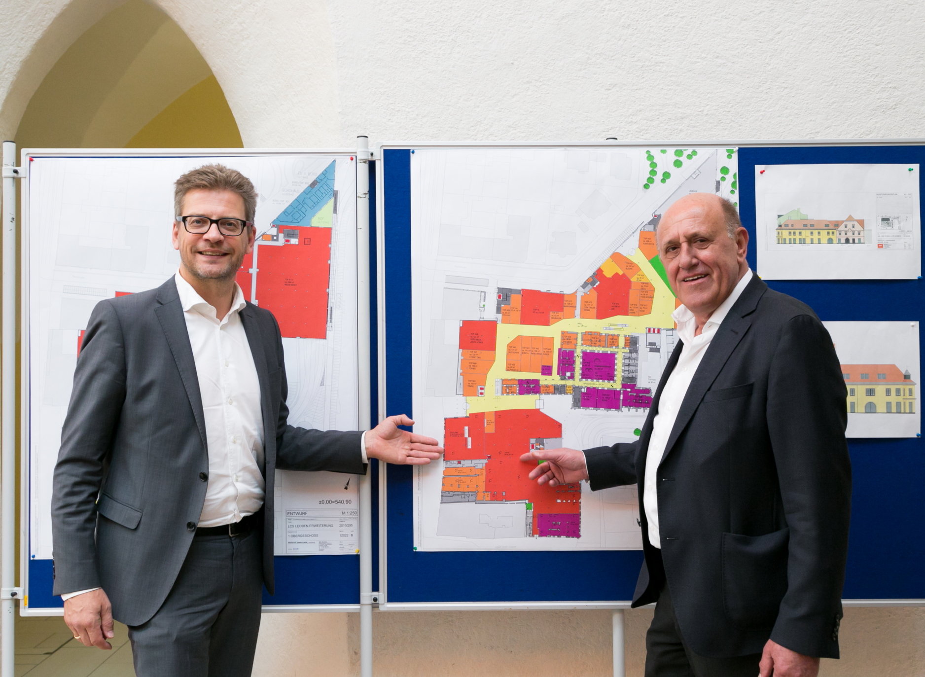 Zwei Männer in Anzügen stehen vor einem schwarzen Brett, auf dem bunte Architekturpläne hängen. Sie lächeln beide und gestikulieren in Richtung der Pläne, was darauf hindeutet, dass es sich um eine Diskussion oder Präsentation über die Entwürfe handelt.