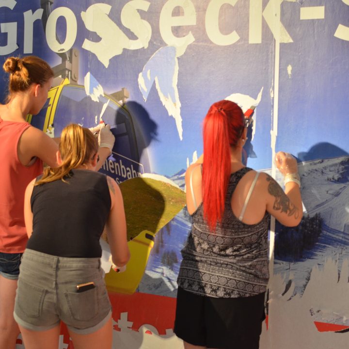 Vier Personen ziehen eine große Werbung von einer Wand ab und geben darunter ein Bild eines schneebedeckten Berges frei. Sie arbeiten gemeinsam unter hellem Licht, wobei in manchen Abschnitten noch Reste des Originalplakats sichtbar sind.