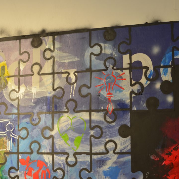 Ein großes Wandbild, das ein buntes Puzzle darstellt, wobei jedes Teil unterschiedliche abstrakte Muster aufweist, darunter eine rote Glühbirne, verschiedene Symbole und helle Farbspritzer. Der Hintergrund ist eine Mischung aus Blau und Schwarz.
