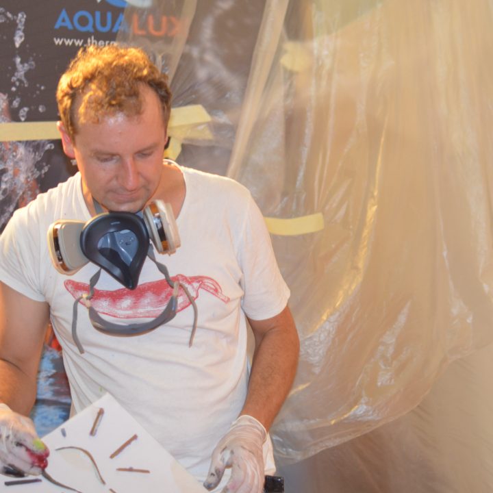 Ein Mann trägt ein weißes T-Shirt mit Krabbenmotiv und eine Atemschutzmaske und hält eine Schablone in der Hand. Er steht vor einer mit Plastik abgedeckten Fläche und bereitet sich wahrscheinlich auf ein Mal- oder Sprühprojekt vor. Im Hintergrund sind verschiedene Werkzeuge und Materialien zu sehen.