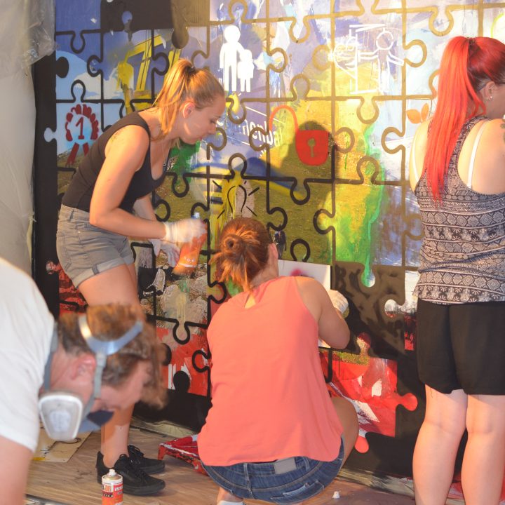 Menschen malen gemeinsam ein farbenfrohes Wandbild aus großen Puzzleteilen an eine Wand. Sie verwenden verschiedene Maltechniken, darunter Sprühfarbe und Pinsel, und arbeiten in einer entspannten Innenumgebung.