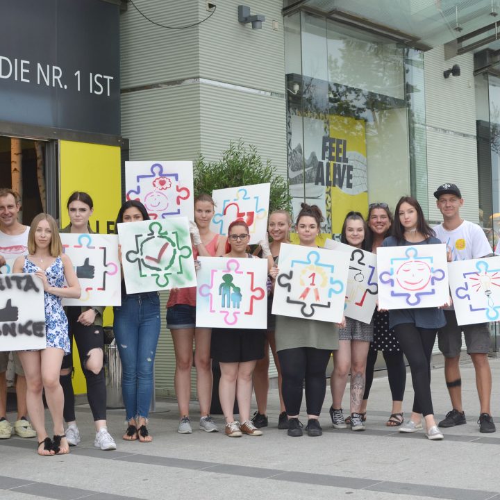 Eine Gruppe von Menschen steht vor einem Gebäude. Jeder hält ein Schild mit einem bunten Puzzleteil-Symbol. Die Gruppe scheint zu lächeln. Im Hintergrund sind Pflanzen und Glasfenster zu sehen.