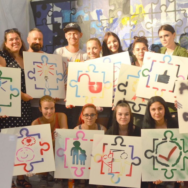 Eine Gruppe lächelnder Menschen hält ein Puzzle-Kunstwerk in der Hand, jedes mit einem anderen Motiv, darunter ein Daumen nach oben, Silhouetten und Symbole. Sie posieren gemeinsam im Innenbereich und drücken so Kreativität und Teamwork aus.