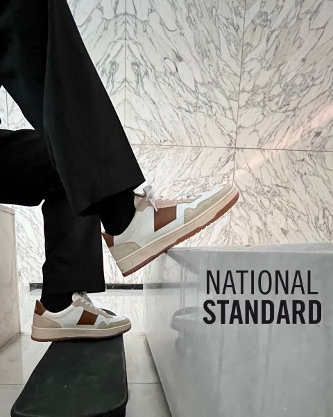 Eine Person in weißen und braunen Turnschuhen steigt vor einer Marmorwand auf eine Treppe. An der Wand steht „NATIONAL STANDARD“. Die Person trägt eine schwarze Hose.