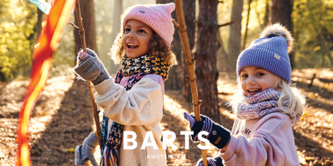 Zwei Kinder laufen durch einen sonnenbeschienenen Wald. Sie tragen Wintermützen, Schals und Handschuhe. Sie lächeln und halten Stöcke mit bunten Bändern in der Hand. Unten im Bild ist der Text „BARTS AMSTERDAM“ zu sehen.