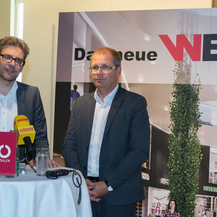 Zwei Männer in Anzügen stehen an einem kleinen runden Tisch mit Mikrofonen und sprechen bei einer Veranstaltung. Im Hintergrund ist „Das neue WEZ“ mit Bildern des Interieurs eines Einkaufszentrums zu sehen. Die Kulisse scheint formell zu sein, möglicherweise eine Pressekonferenz oder Präsentation.