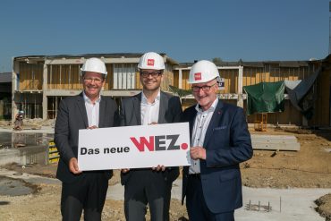 Drei Männer in Anzügen und Bauhelmen stehen lächelnd auf einer Baustelle und halten ein Schild mit der Aufschrift „Das neue WEZ“. Hinter ihnen ist ein halbfertiges Gebäude unter einem strahlend blauen Himmel zu sehen.