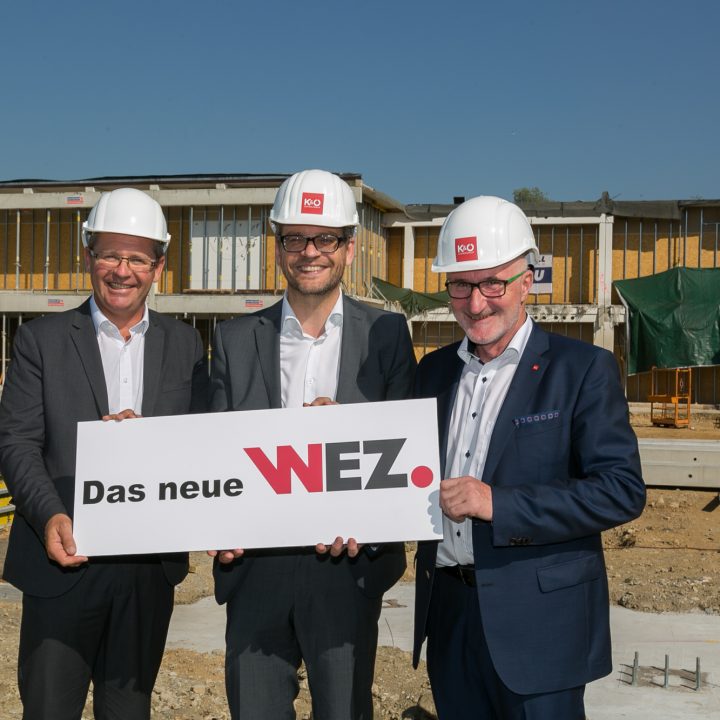 Drei Männer in Anzügen und Bauhelmen stehen lächelnd auf einer Baustelle und halten ein Schild mit der Aufschrift „Das neue WEZ“. Hinter ihnen ist ein halbfertiges Gebäude unter einem strahlend blauen Himmel zu sehen.
