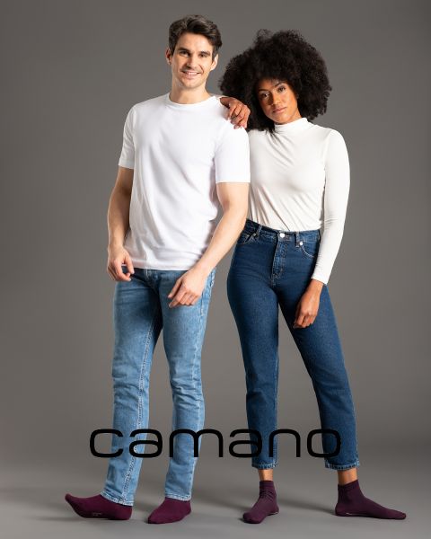 Ein Mann in weißem T-Shirt und Jeans und eine Frau in langärmligem weißem Oberteil und Jeans posieren mit den Armen umeinander. Beide sind barfuß. Unten im Bild steht das Wort „camano“.