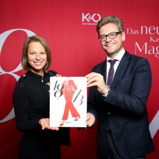 Zwei Menschen lächeln und halten eine Zeitschrift vor einem roten Hintergrund mit dem Text „K&Ö“ und „Das neue K&Ö Magazin“. Der Mann trägt Brille und Anzug, die Frau ein schwarzes Outfit.