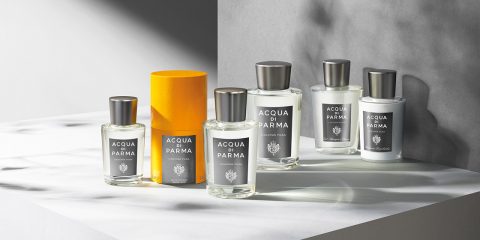 Flaschen Acqua di Parma Cologne werden auf einer grauen Oberfläche mit weichen Schatten ausgestellt. Eine Flasche befindet sich in einer orangefarbenen zylindrischen Schachtel, während die anderen um sie herum angeordnet sind und alle silberne Verschlüsse und Etiketten aufweisen.