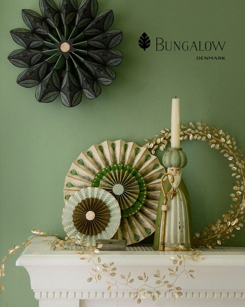 Eine grüne Wand ist mit Dekorationsgegenständen geschmückt: ein schwarzes sternförmiges Ornament, runde Papierfächer in Grün und Beige, eine rustikale Figur einer Person in einer Robe mit hohem Hut, ein goldener dekorativer Kranz und ein weißer Kaminsims mit Blumenmustern. Der Text lautet „Bungalow Dänemark“.