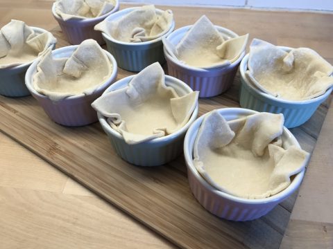 Ungebackene Tortenböden werden in kleine, bunte Keramikförmchen auf einer Holzoberfläche gelegt. Die Förmchen werden in einer ordentlichen Reihe aufgestellt und sind bereit zum Befüllen und Backen.